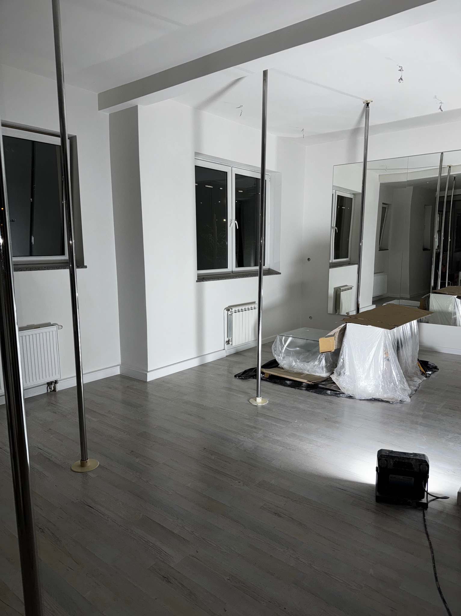 Białe wnętrze z zamontowanymi drążkami do ćwiczeń, lustrami i podłogą imitującą drewno. Meble w folii ochronnej. Oświetlenie robocze na podłodze.
