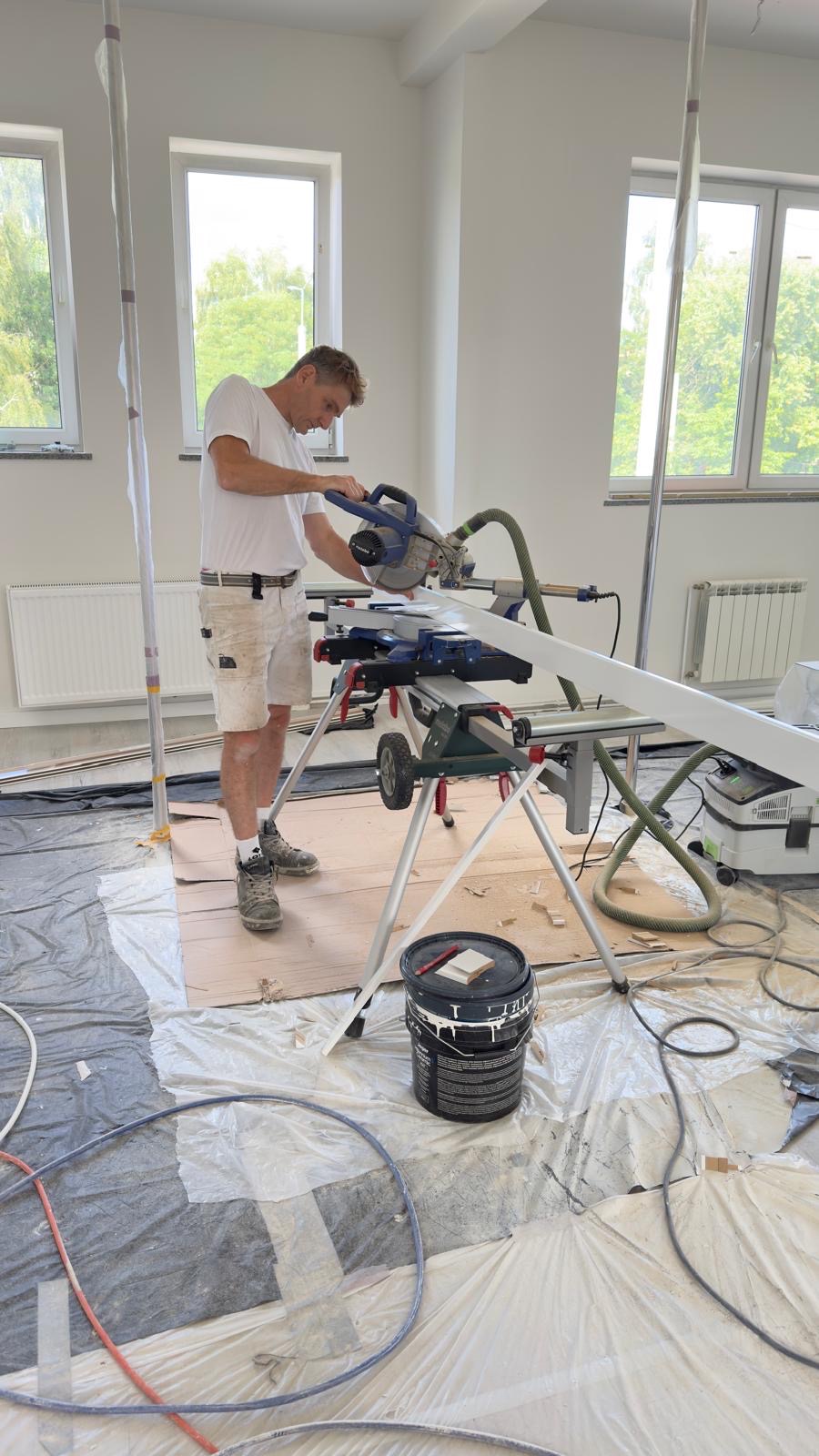Mężczyzna przycina białą listwę przypodłogową piłą elektryczną w jasnym pokoju, przygotowanym do prac wykończeniowych. Podłoga zabezpieczona folią.
