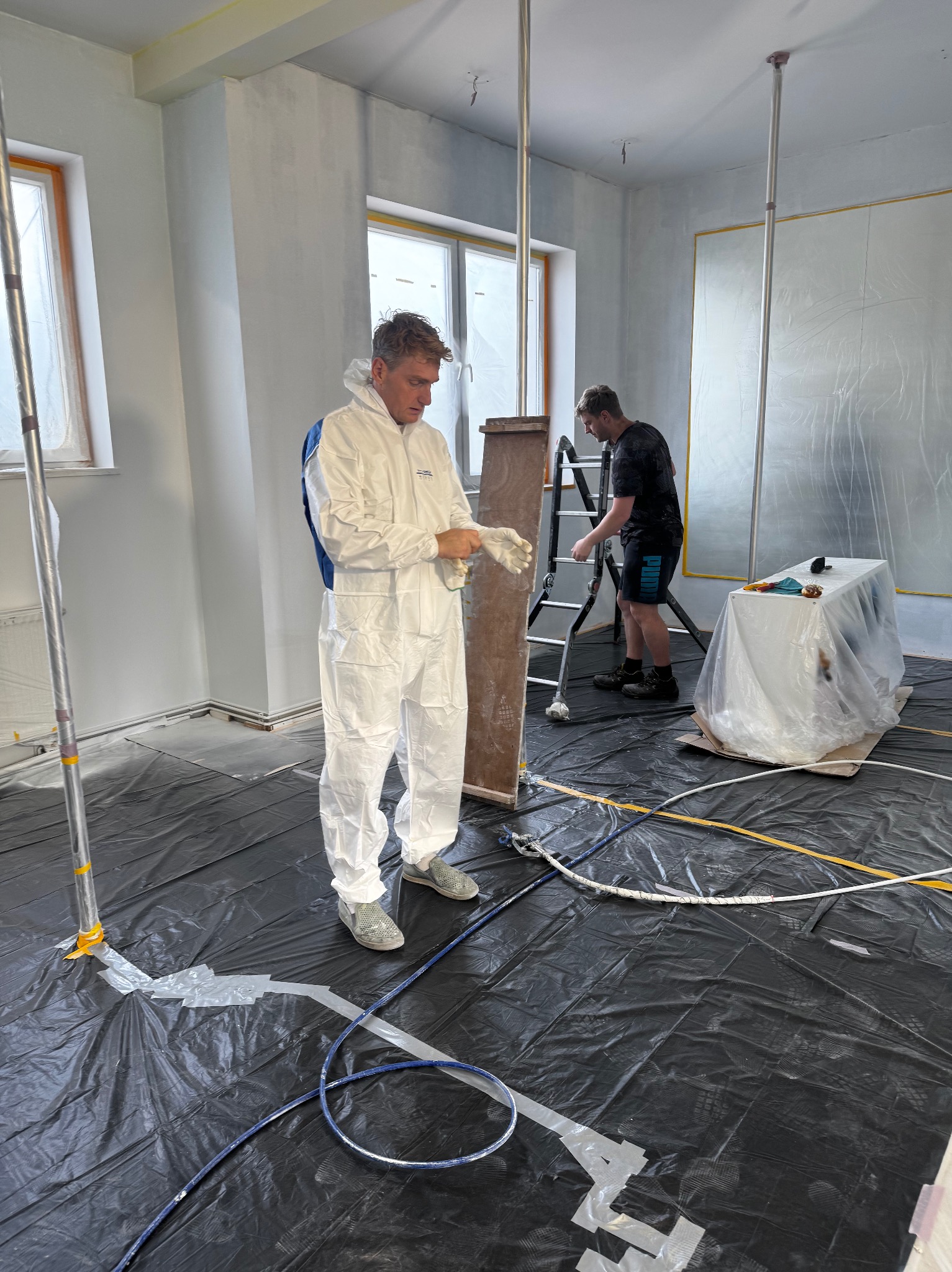 Przygotowania do malowania wnętrza: pracownik w kombinezonie zakłada rękawice, drugi maluje ściany z drabiny. Podłoga zabezpieczona folią.