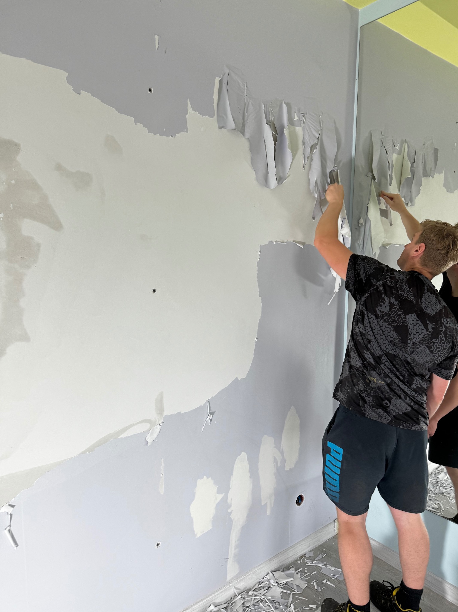 Usuwanie starej tapety ze ściany w pokoju. Widoczny fragment ściany z resztkami kleju i odchodzącej tapety, oraz mężczyzna z narzędziem w trakcie pracy.