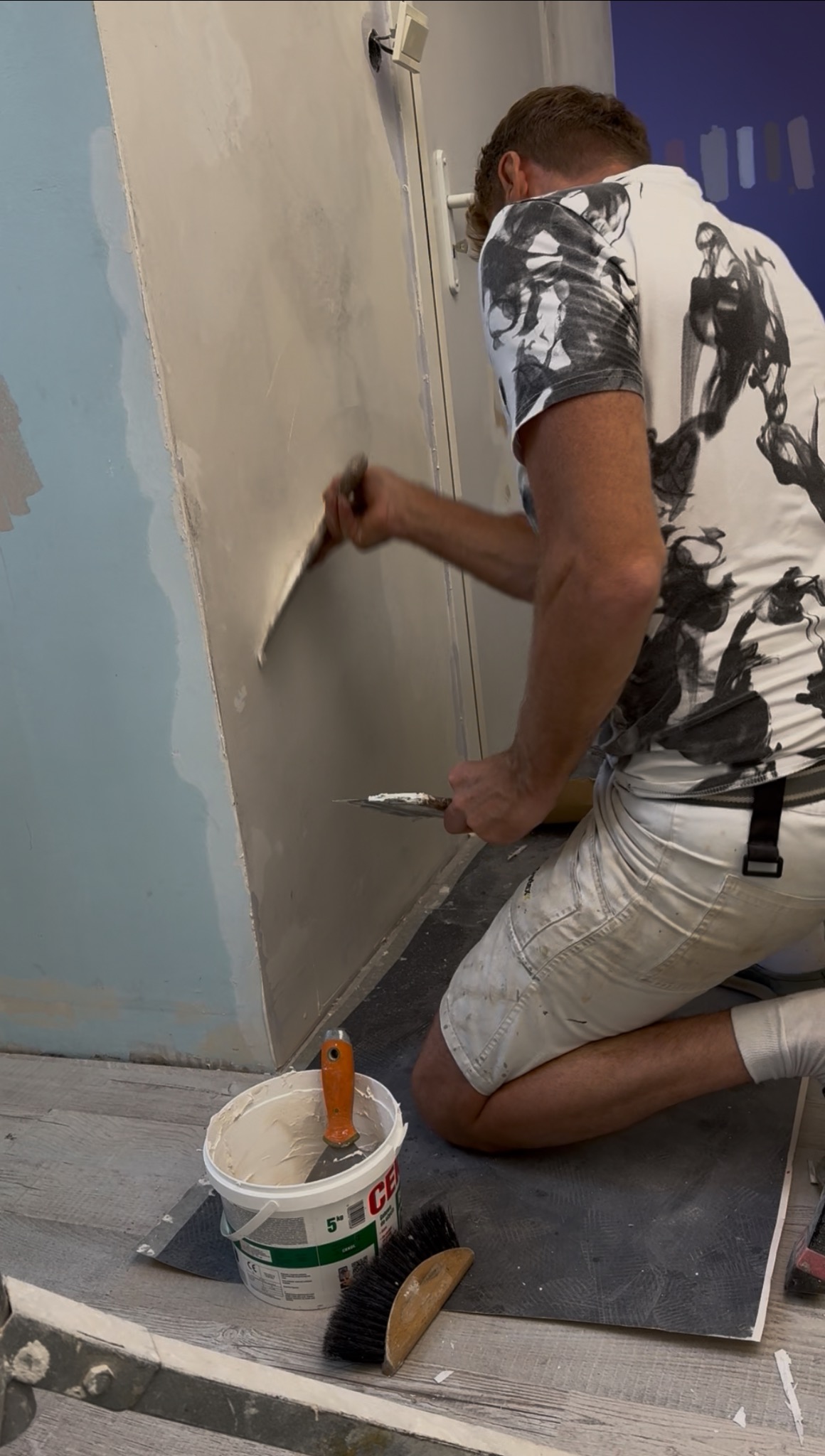 Pracownik szpachluje ścianę w pomieszczeniu. Widoczny kubeł z masą szpachlową, szpachelka i pędzel. Na ścianie ślady po wcześniejszych pracach.