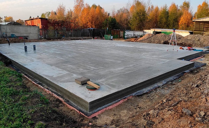 Wylany, świeży betonowy fundament płyty z izolacją pionową, widoczne rury kanalizacyjne i niwelator laserowy na statywie w tle.