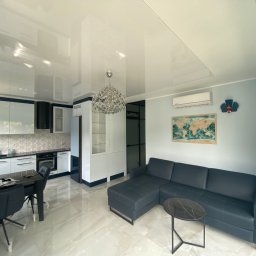 Katarzyna Putynkowska - Nowoczesny salon z aneksem kuchennym, błyszczący sufit podwieszany, szary narożnik, okrągły stolik kawowy z marmurowym blatem, mapa świata na ścianie.