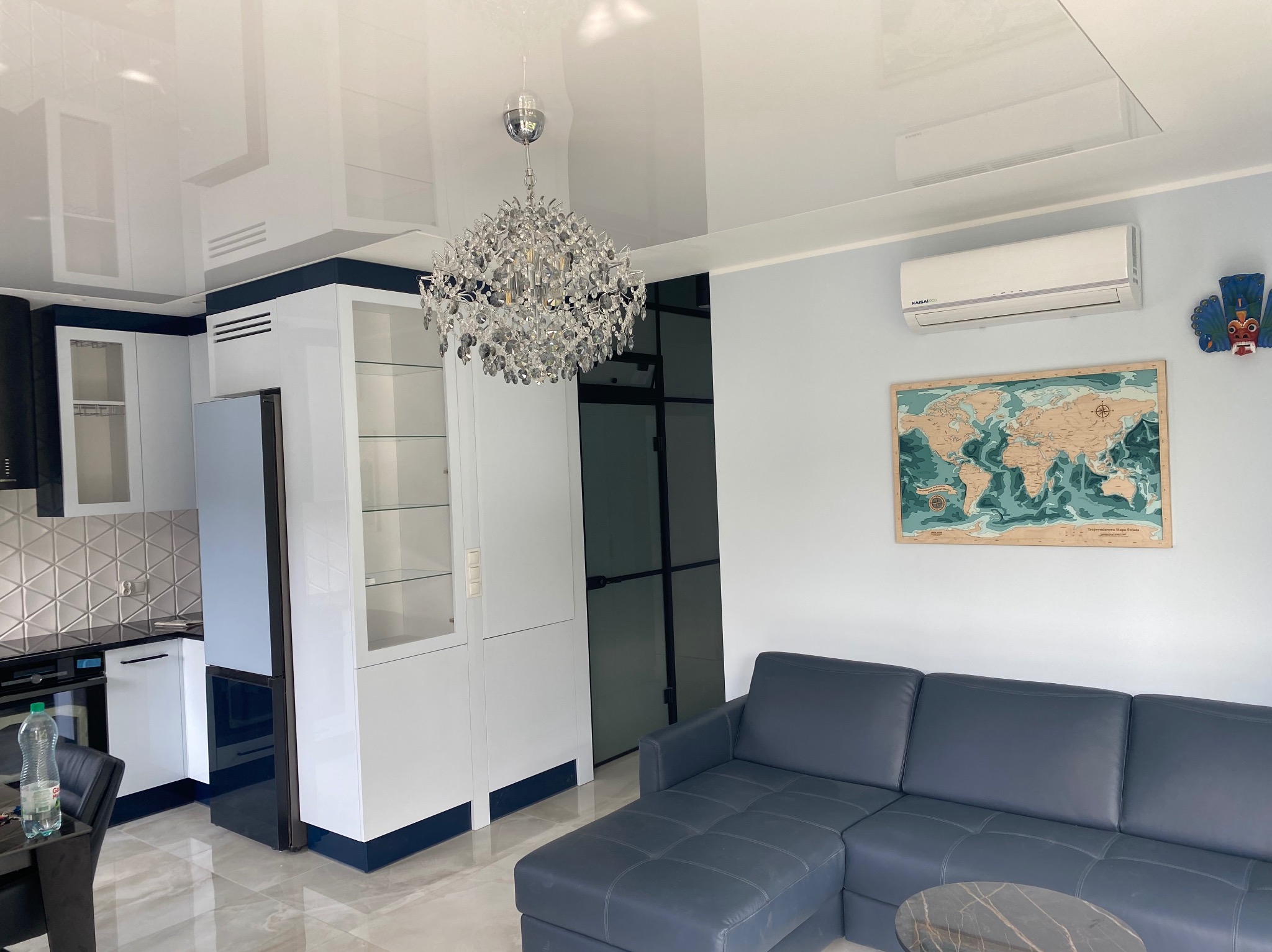 Nowoczesne wnętrze salonu z aneksem kuchennym w jasnych kolorach, z błyszczącym sufitem, kryształowym żyrandolem i szarą sofą narożną.