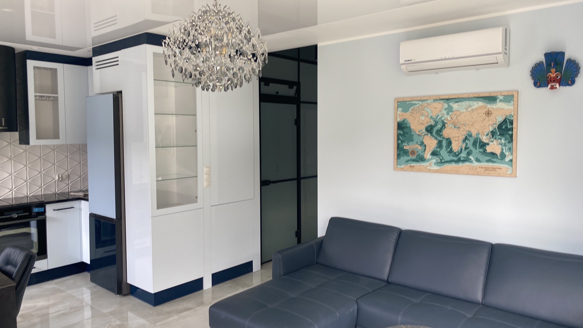Nowoczesne wnętrze mieszkania z aneksem kuchennym, białymi meblami z granatowymi akcentami, kryształowym żyrandolem i szarą sofą narożną, uzupełnione mapą świata na ścianie.