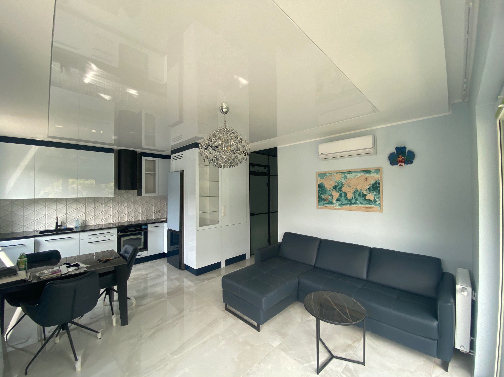 Nowoczesny salon z aneksem kuchennym, błyszczący sufit podwieszany, szary narożnik, okrągły stolik kawowy z marmurowym blatem, mapa świata na ścianie.