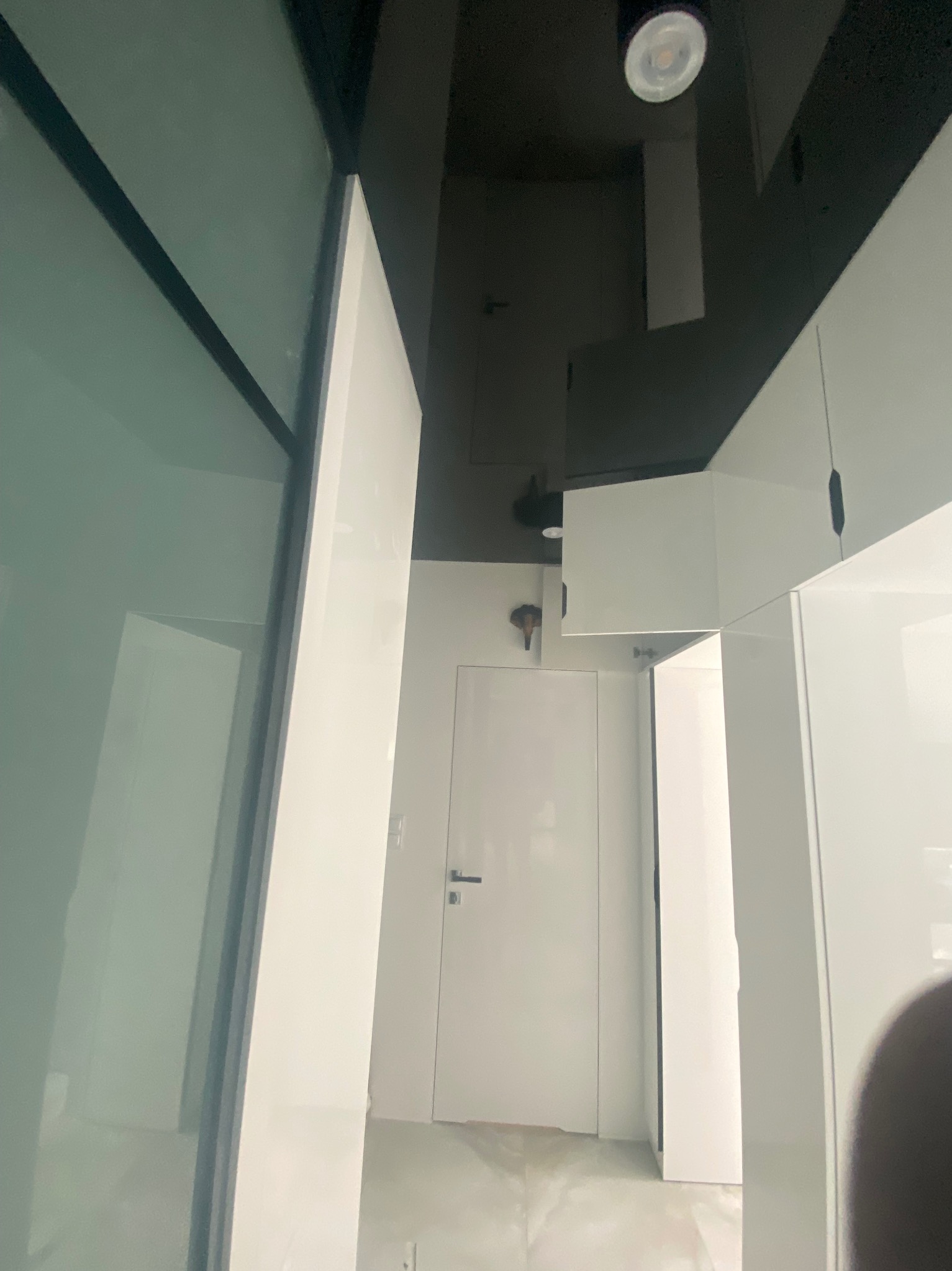 Nowoczesne wnętrze z białymi, lakierowanymi szafkami pod sufitem i czarnym sufitem podwieszanym z oświetleniem punktowym, widok na białe drzwi wewnętrzne.