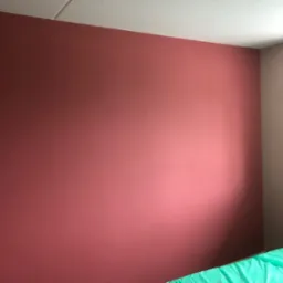 Ściana w pokoju pomalowana na kolor terakoty, sąsiadująca z beżową ścianą przy oknie, widoczna część łóżka z zieloną narzutą.