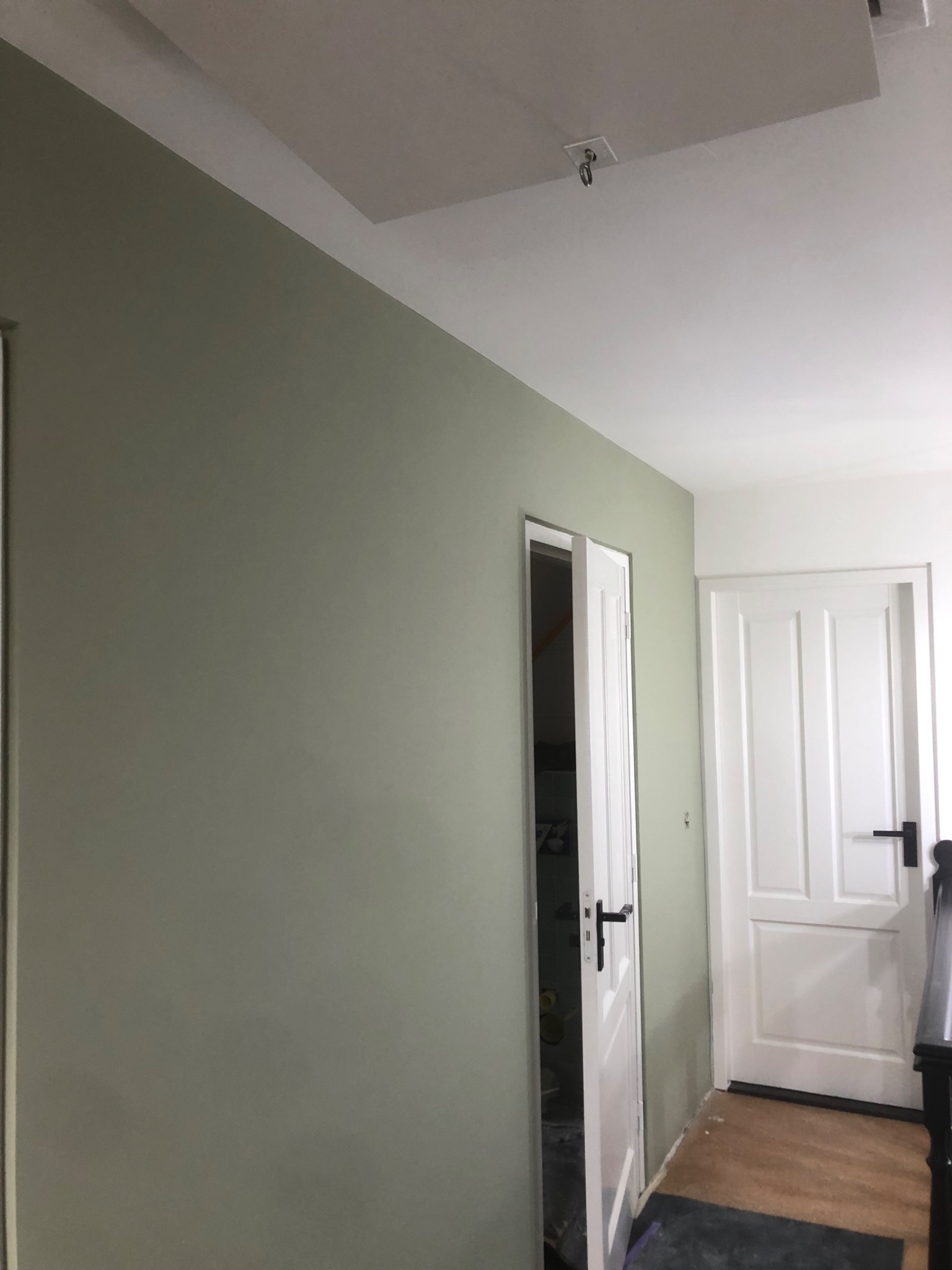 Świeżo pomalowana ściana w odcieniu pistacjowej zieleni w korytarzu, widoczne dwie białe drzwi z czarnymi klamkami, sufit biały z widocznym włazem.