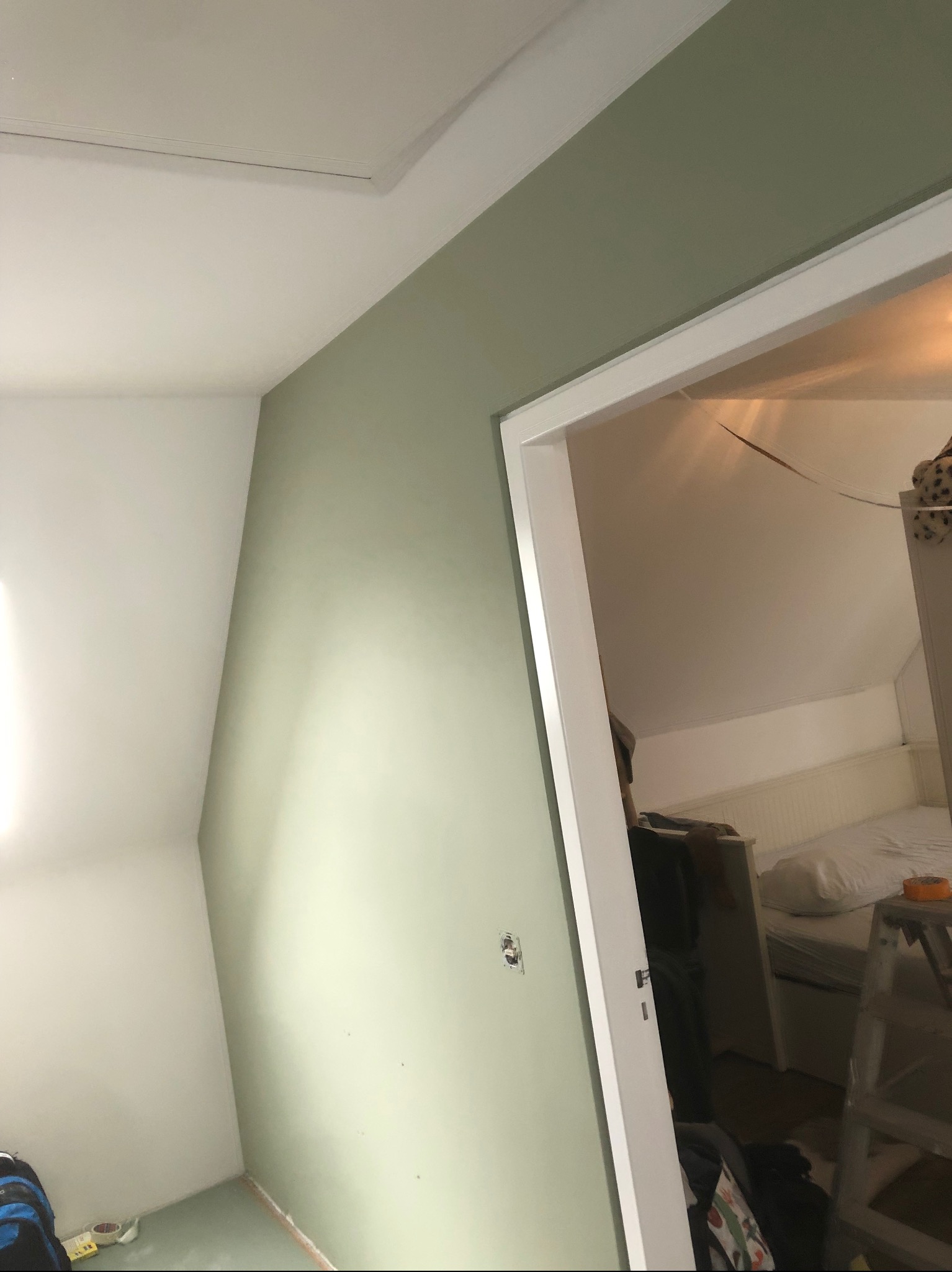 Świeżo pomalowana ściana w pokoju na poddaszu w odcieniach zieleni, widoczne gniazdko elektryczne, fragment sufitu i futryna drzwi prowadząca do pokoju z łóżkiem i drabiną.