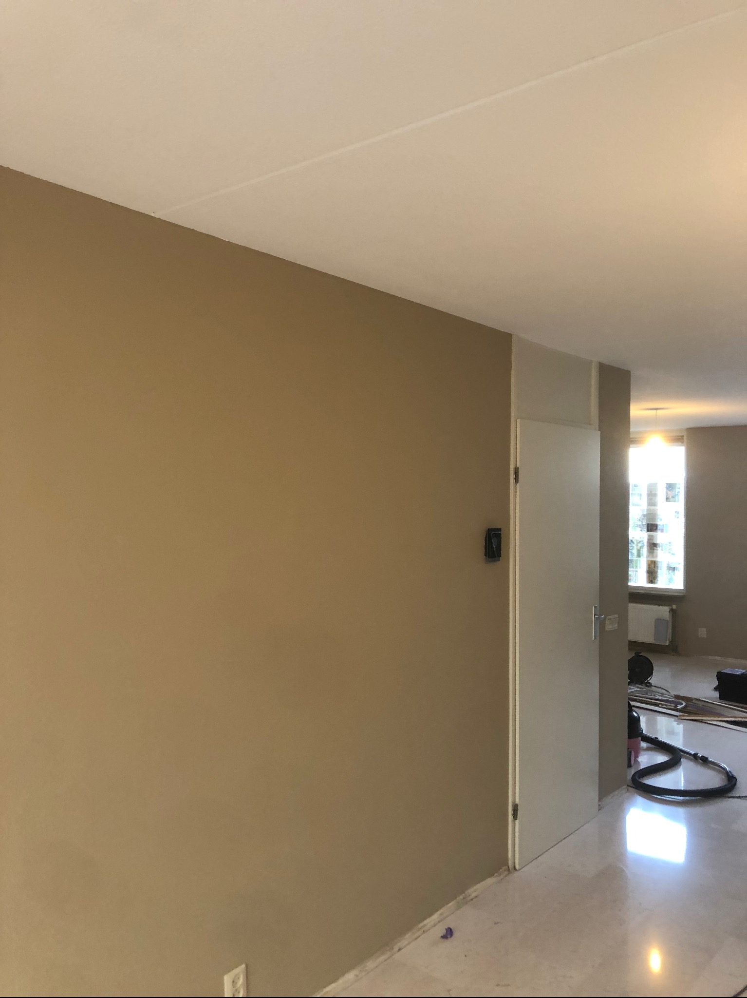 Świeżo pomalowane ściany w odcieniach beżu i bieli w jasnym mieszkaniu, widoczna gładka powierzchnia ścian, drzwi i fragment podłogi z połyskiem, w tle okno z widokiem na zewnątrz i sprzęt malarski.