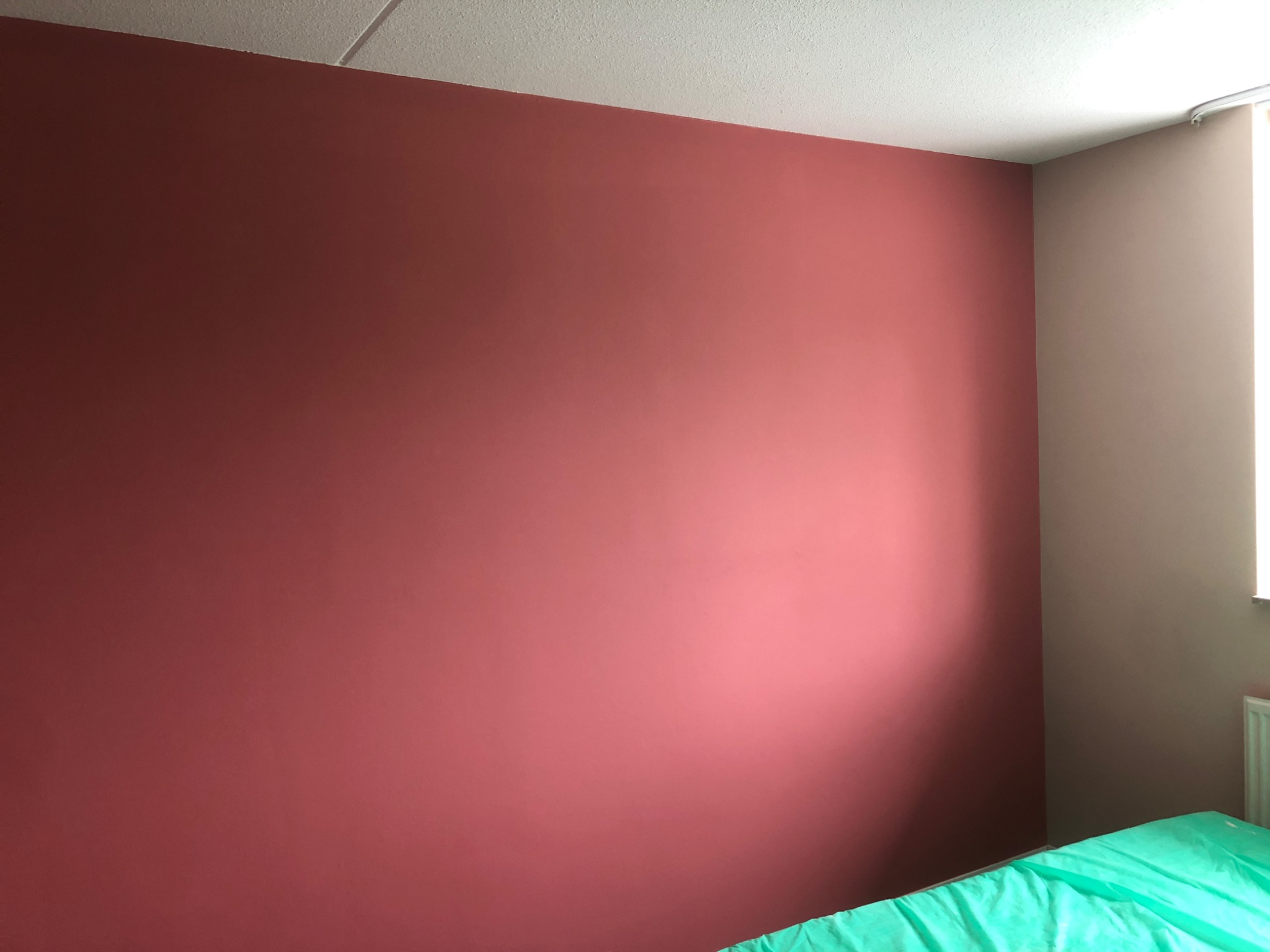 Ściana w pokoju pomalowana na kolor terakoty, sąsiadująca z beżową ścianą przy oknie, widoczna część łóżka z zieloną narzutą.