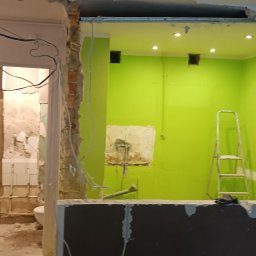 Sebastian Kuźmiński - Widok na remontowaną łazienkę z fragmentem toalety, przez otwór w ścianie z widoczną konstrukcją z płyty karton gips, z przewodami elektrycznymi i fragmentami starej glazury oraz drabina w tle...