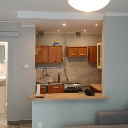 Sebastian Kuźmiński - Widok na odnowione wnętrze mieszkania z otwartą kuchnią i łazienką z kabiną prysznicową, ściany pomalowane na szaro, drewniane szafki kuchenne i biała armatura łazienkowa.