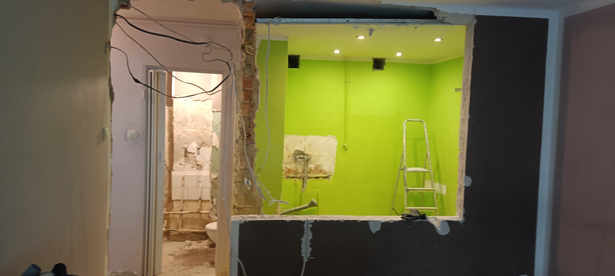 Widok na remontowaną łazienkę z fragmentem toalety, przez otwór w ścianie z widoczną konstrukcją z płyty karton gips, z przewodami elektrycznymi i fragmentami starej glazury oraz drabina w tle...