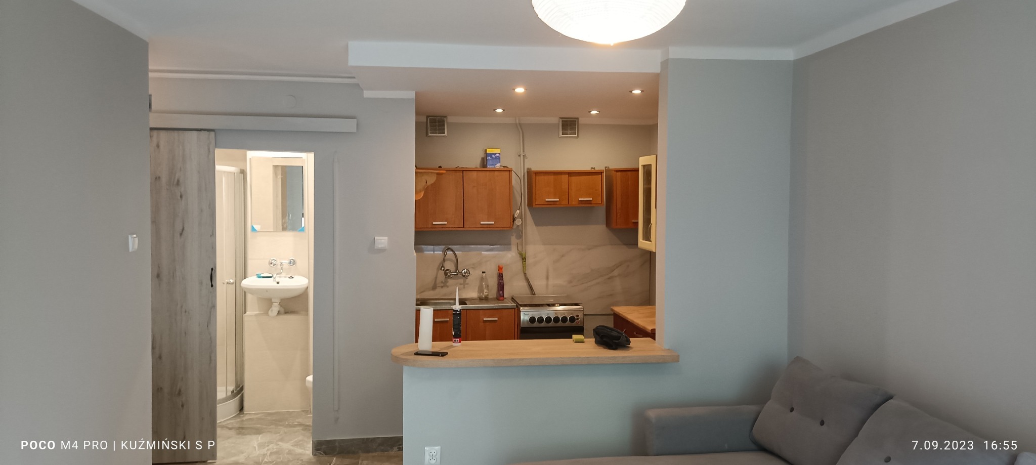 Widok na odnowione wnętrze mieszkania z otwartą kuchnią i łazienką z kabiną prysznicową, ściany pomalowane na szaro, drewniane szafki kuchenne i biała armatura łazienkowa.