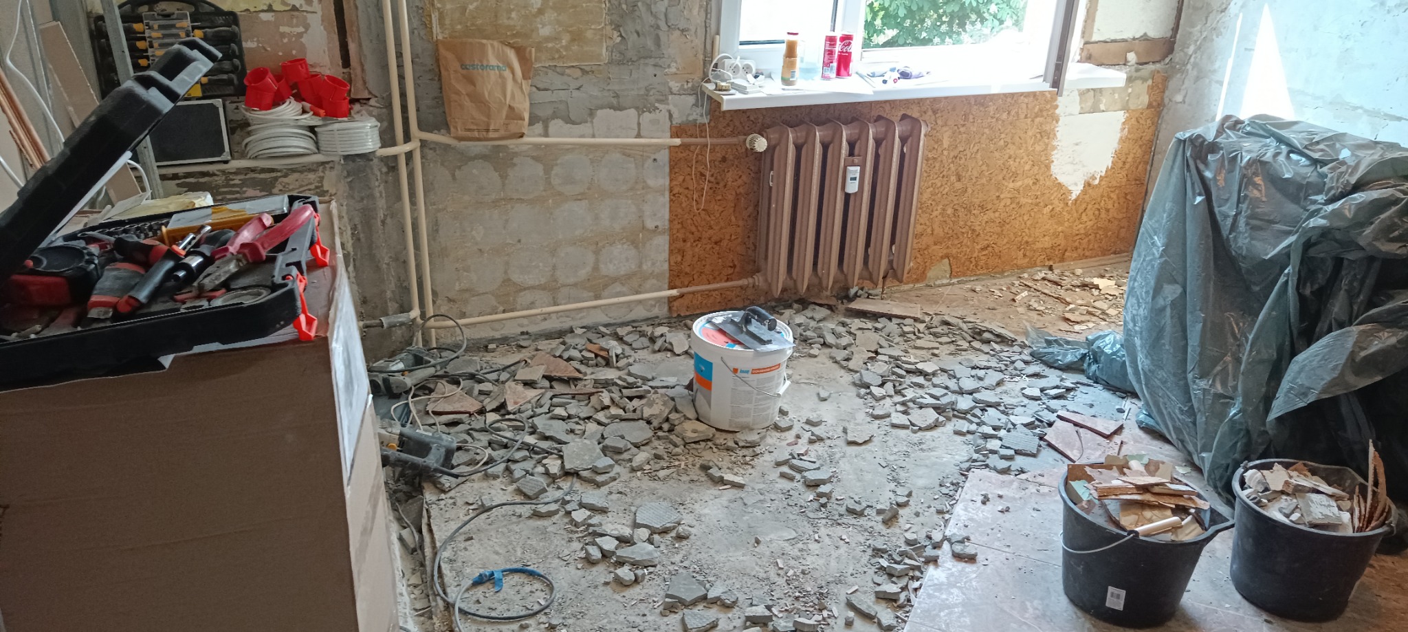 Fragment pokoju w trakcie prac przygotowawczych do położenia płytek: gruz po skuciu starej glazury na podłodze, wiadro z zaprawą, narzędzia w skrzynce, odsłonięte rury, okno z parapetem.