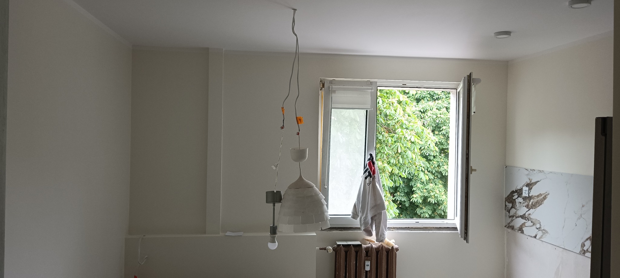 Pomalowane na biało wnętrze pokoju z wiszącą lampą bez klosza, otwartym oknem z widokiem na zieleń i grzejnikiem centralnego ogrzewania pod oknem, z widocznymi śladami prac wykończeniowych na ścianie.