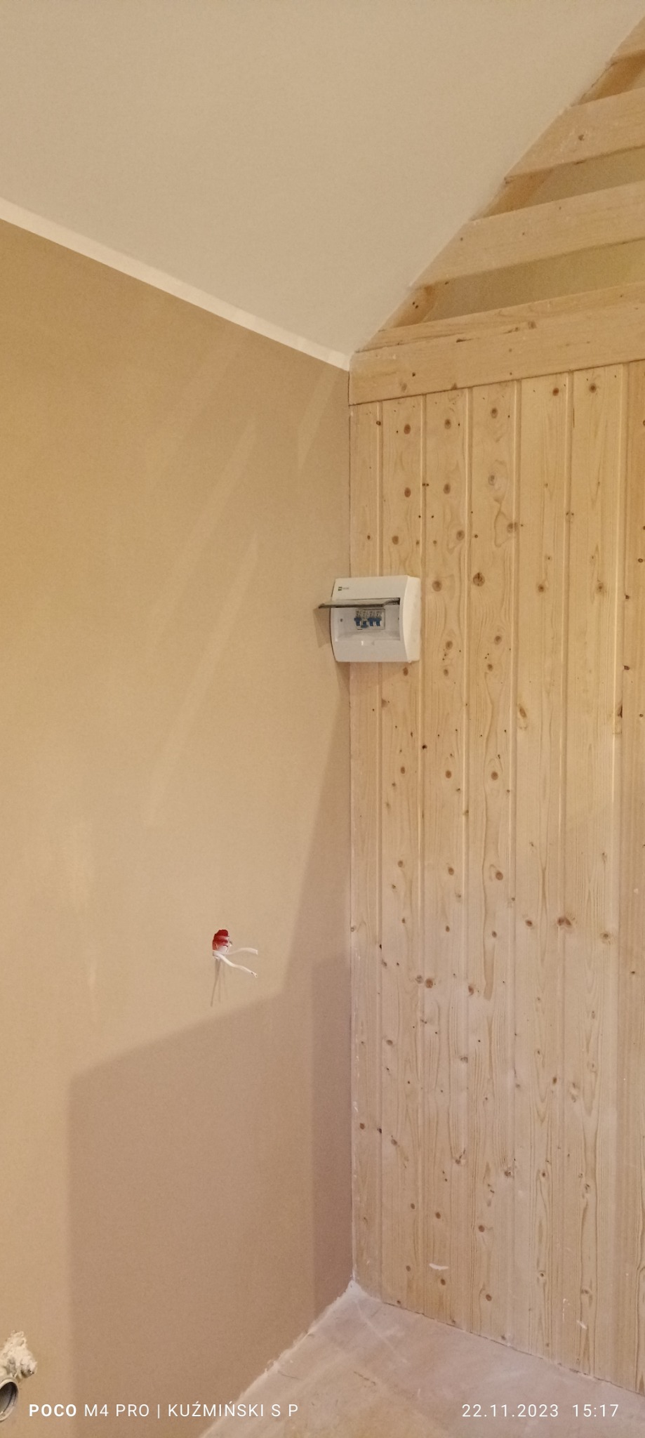 Wnętrze w trakcie wykańczania: ściana szpachlowana w beżowym odcieniu, drewniana boazeria pionowa, puszka elektryczna na ścianie i wystające kable ze ściany.