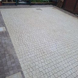Marcin Stępień - Jasnoszara kostka granitowa ułożona na podjeździe, widoczna studzienka kanalizacyjna, obrzeża z ciemniejszej kostki.