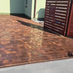 Ułożona, brązowa kostka brukowa przed domem z drewnianym ogrodzeniem, częściowo mokra po myciu, z widocznym cieniem osoby.