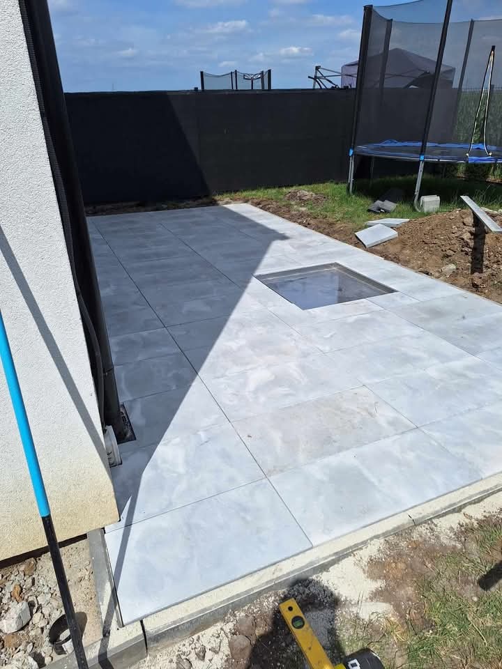 Jasnoszara, kwadratowa nawierzchnia z płyt betonowych na zewnątrz budynku, z widocznym otworem na studzienkę rewizyjną i poziomnicą w rogu. W tle trampolina i czarny płot.