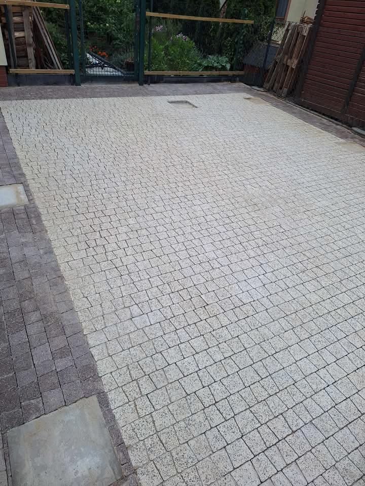 Jasnoszara kostka granitowa ułożona na podjeździe, widoczna studzienka kanalizacyjna, obrzeża z ciemniejszej kostki.