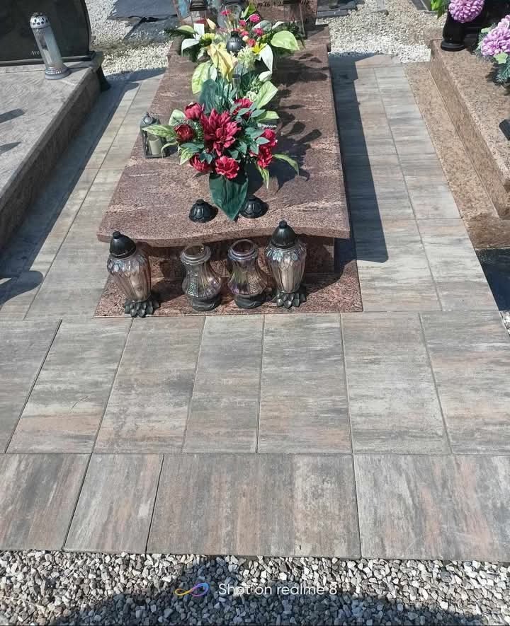 Ułożona kostka brukowa wokół nagrobka z kwiatami i lampionami, wykończona drobnym żwirem.