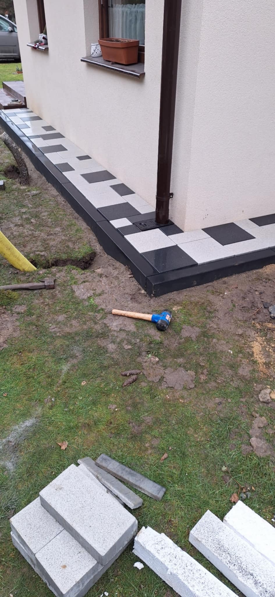Ułożona szachownica z jasnych i ciemnych płyt chodnikowych przy ścianie budynku z rynną, obok leży gumowy młotek brukarski i stos płyt do dalszej pracy.