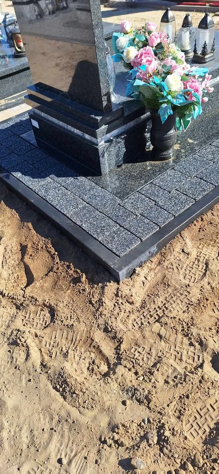 Granitowa kostka brukowa wokół czarnego pomnika z kwiatami, widoczne ślady butów w piasku.