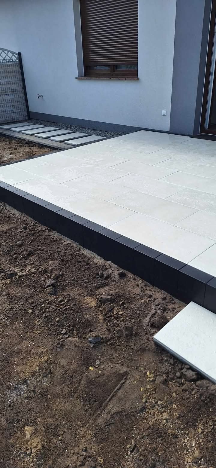 Jasne płyty tarasowe z ciemną obwódką na przygotowanym podłożu ziemnym, widoczne także płyty chodnikowe prowadzące do budynku.