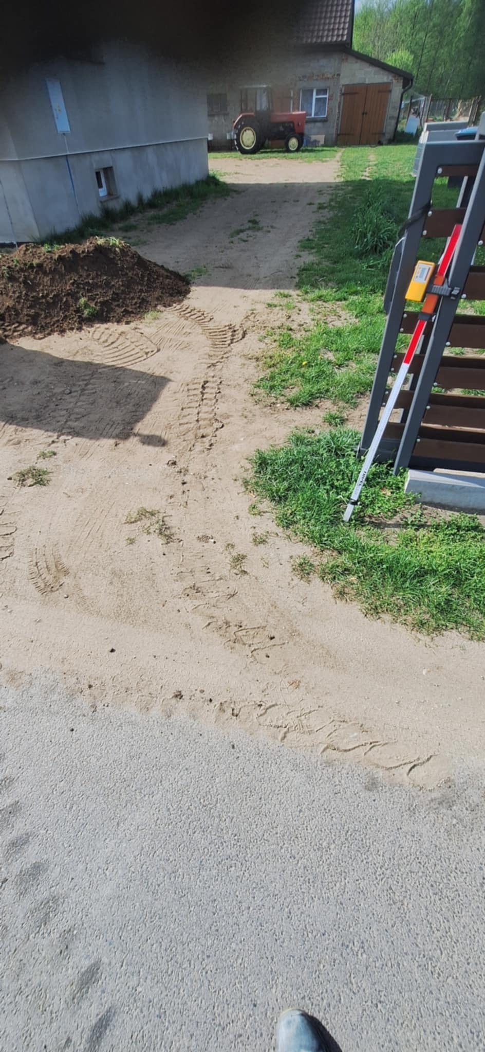Teren po wstępnym wyrównaniu pod utwardzenie drogi, widoczne ślady opon, sterta ziemi i budynek gospodarczy w tle.