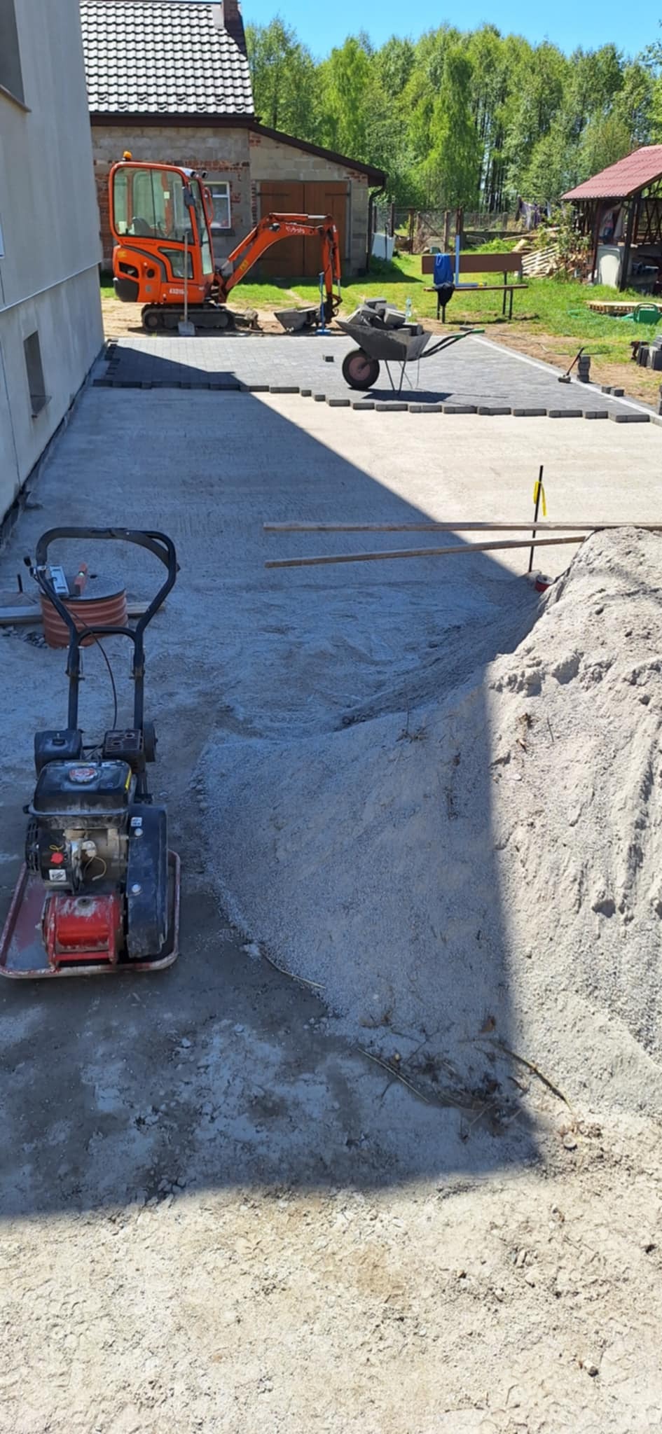 Prace brukarskie w toku: częściowo ułożona kostka, ubijarka wibro, mini koparka i taczka z kostką na posesji. Widoczne przygotowanie terenu pod nawierzchnię.