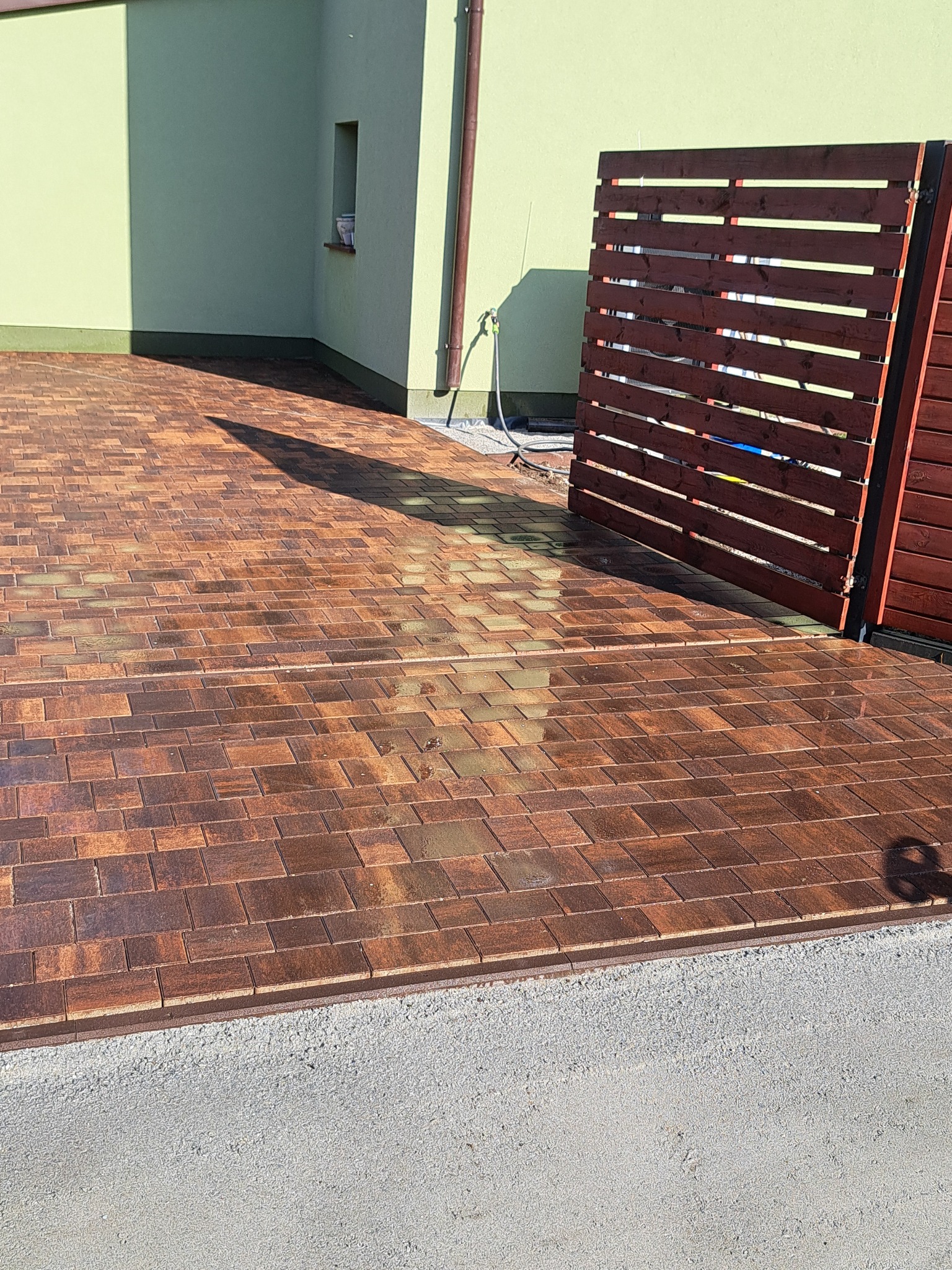 Ułożona, brązowa kostka brukowa przed domem z drewnianym ogrodzeniem, częściowo mokra po myciu, z widocznym cieniem osoby.