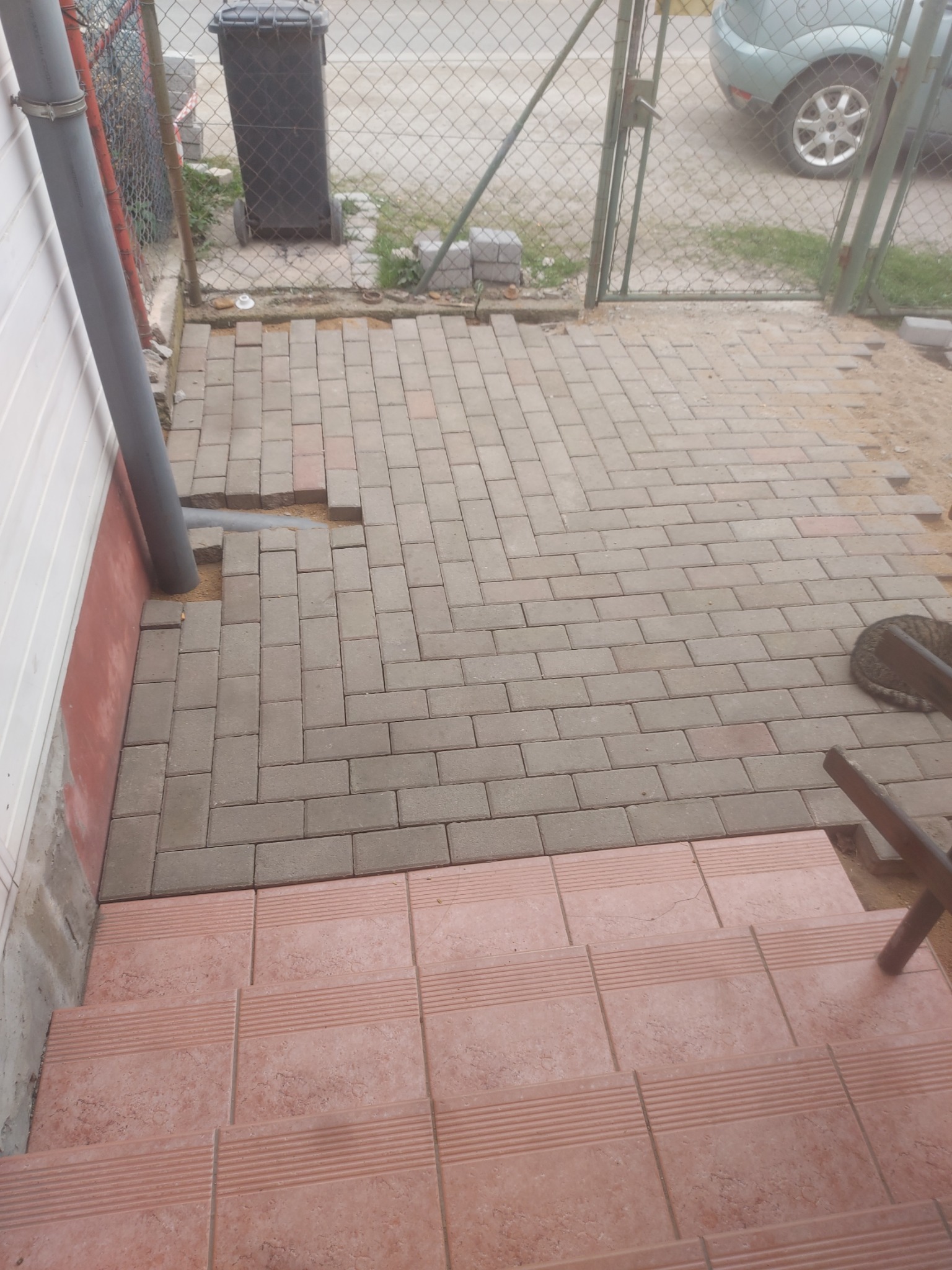 Fragment układanej szarej kostki brukowej w pobliżu ściany budynku z rurą spustową, widoczne różowe płytki schodowe, teren przygotowany pod dalsze układanie, w tle siatka ogrodzeniowa i fragment...