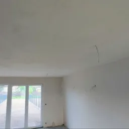 Wnętrze w stanie deweloperskim z wyprowadzonymi przewodami elektrycznymi ze ściany i sufitu, widoczne okno balkonowe z widokiem na zewnątrz.