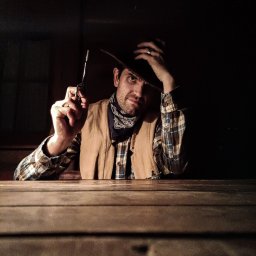 VT Events Fun Magdalena Wojciechowska - Mężczyzna w stroju kowboja, z chustą i kapeluszem, siedzi przy drewnianym stole i celuje z rewolweru. Mroczna sceneria, klimatyczny portret.