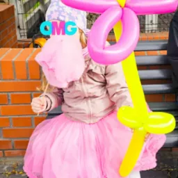 Dziewczynka w różowej tutu trzyma watę cukrową i balon w kształcie kwiatka na tle muru z cegły i zieleni.