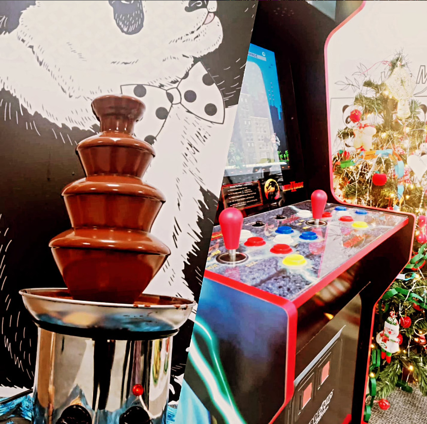 Czekoladowa fontanna na metalowej podstawie, obok automat do gier z joystickami i choinka z ozdobami w tle, całość na tle grafiki z pandą w muszce.