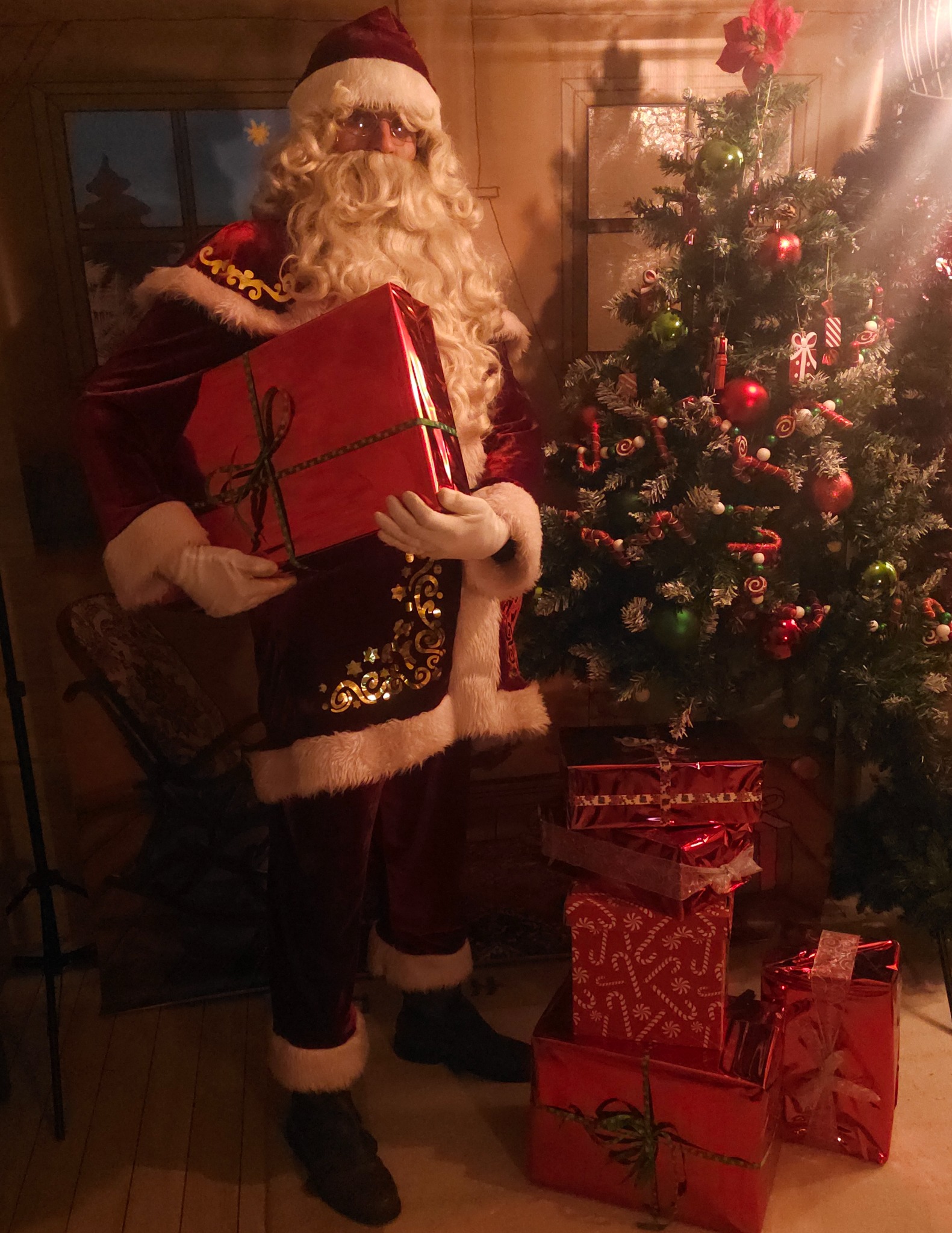 Uśmiechnięty Święty Mikołaj w czerwonym stroju trzyma prezent na tle choinki i stosu prezentów w czerwonych opakowaniach.