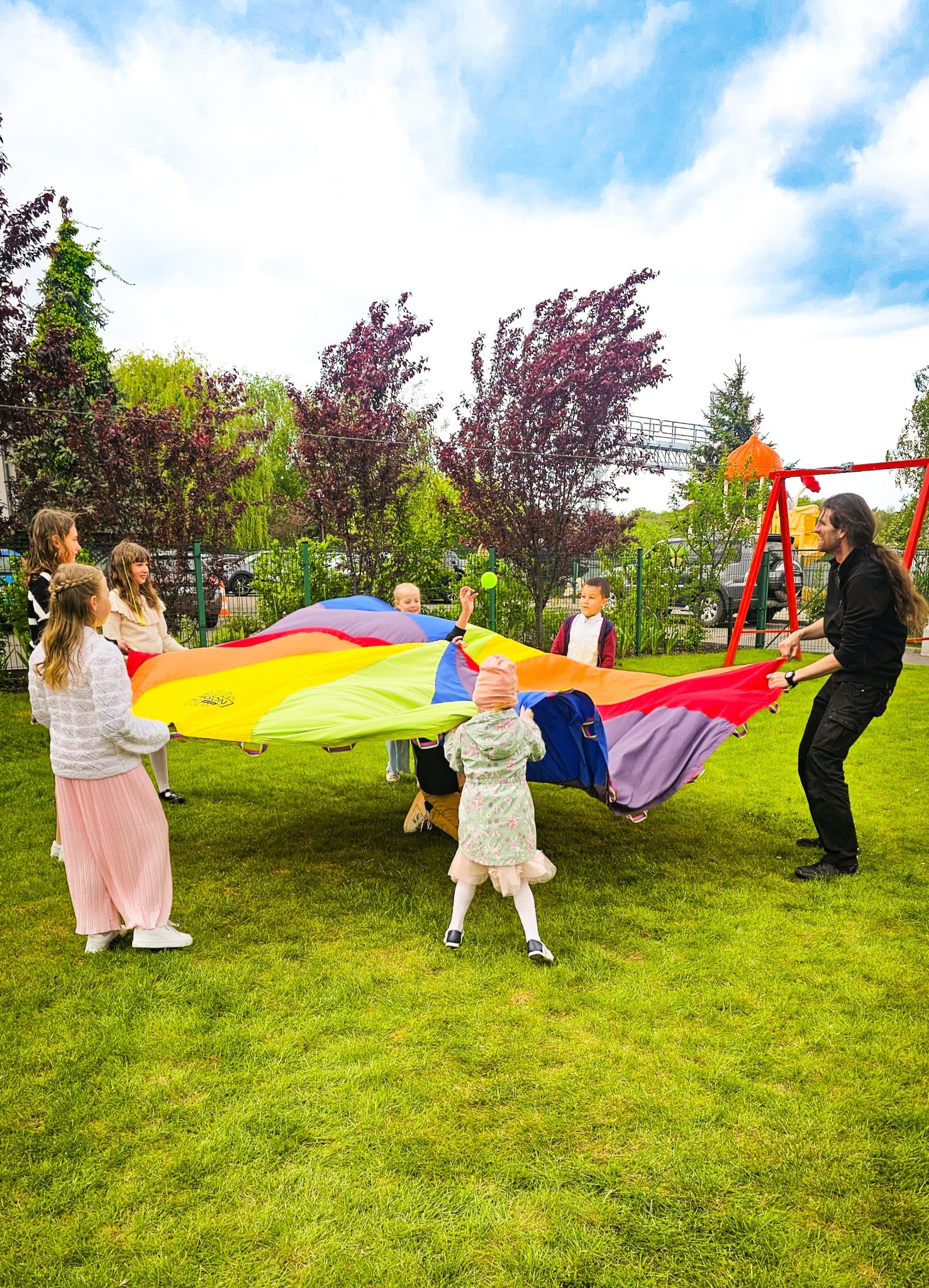 Animator z grupą dzieci bawi się kolorową chustą animacyjną na trawie. W tle drzewa i plac zabaw. Aktywna zabawa na świeżym powietrzu.