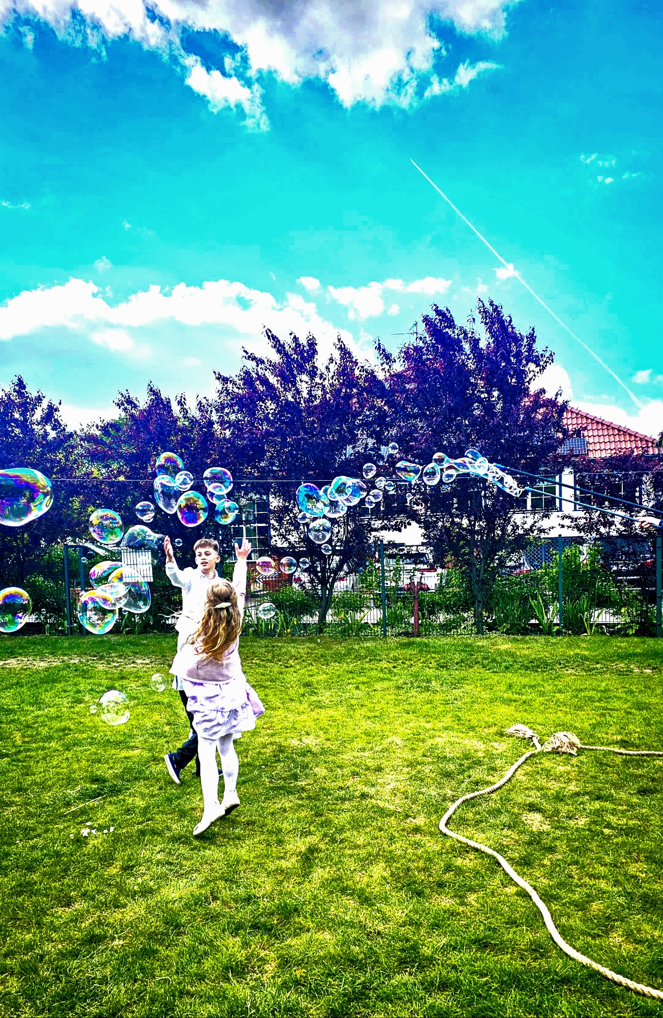 Dzieci łapią bańki mydlane na trawie w słoneczny dzień. Chłopiec w białej koszuli i dziewczynka w fioletowej sukience. W tle drzewa i niebieskie niebo.