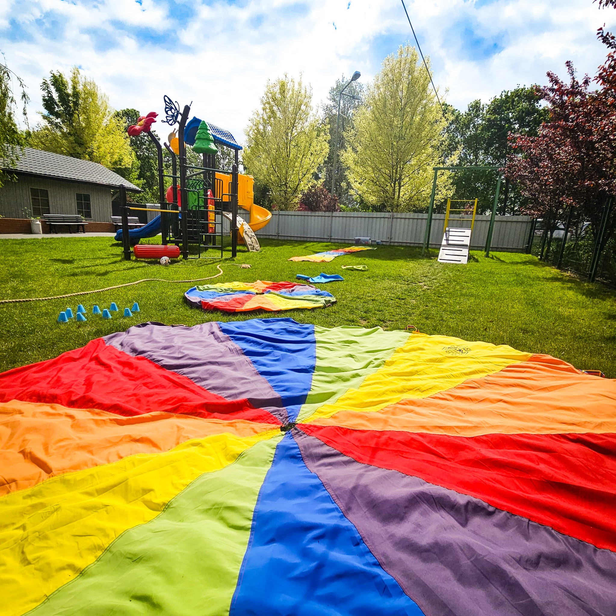 Kolorowa chusta animacyjna rozłożona na trawie placu zabaw z elementami wyposażenia, w tle budynek i drzewa pod błękitnym niebem.