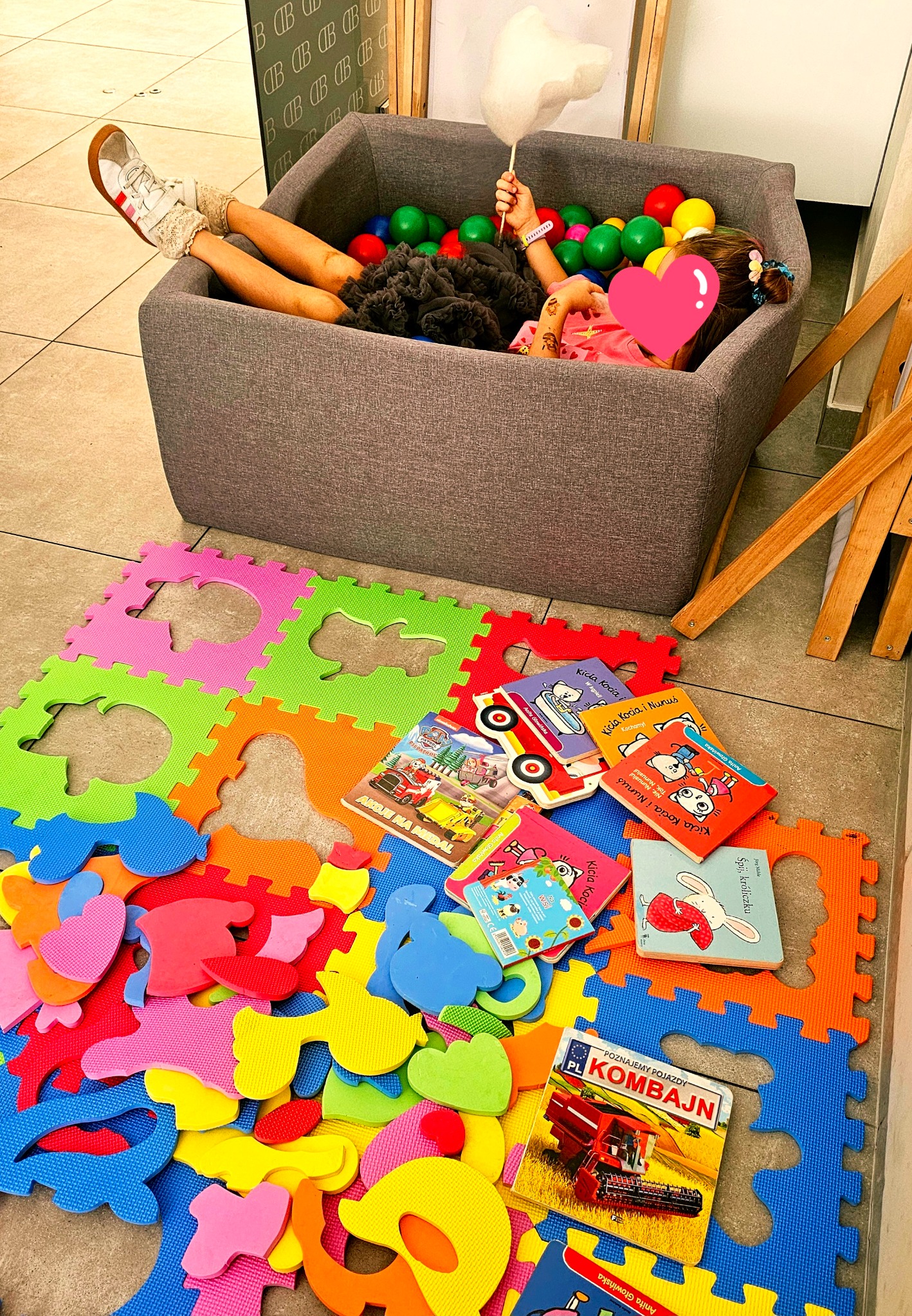 Dziewczynka w basenie z kulkami trzyma watę cukrową. Obok kolorowe puzzle piankowe i książeczki dla dzieci. Aranżacja zabawy dla dzieci, być może w agencji eventowej.