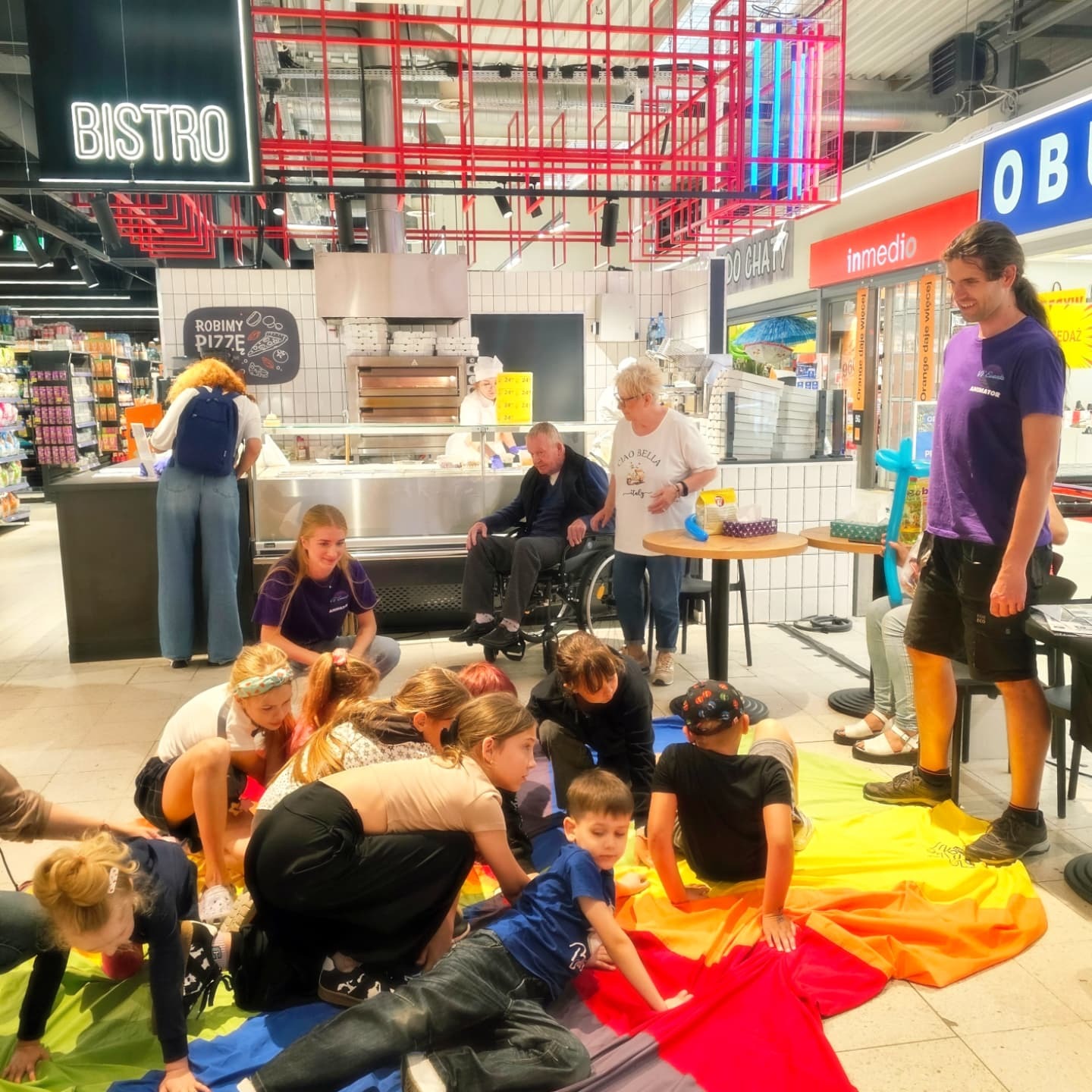 Animator w fioletowej koszulce prowadzi zabawy z dziećmi na kolorowej chuście w supermarkecie, w tle bistro z piecem do pizzy i klienci.