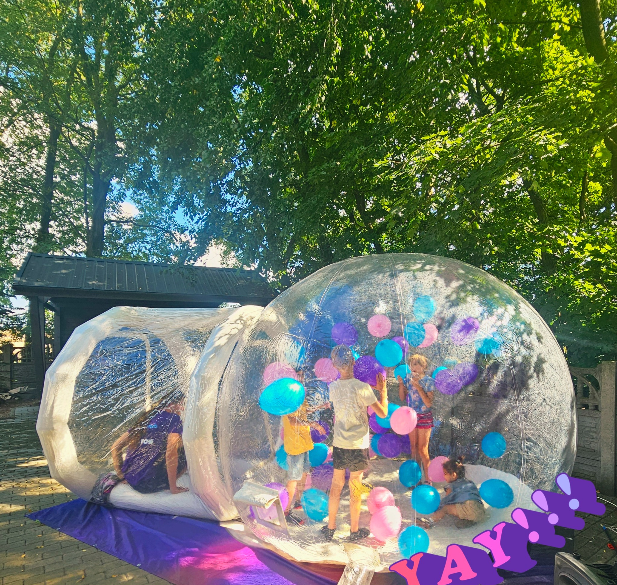 Dzieci wewnątrz dmuchanej kuli z kolorowymi balonami, dekoracja 'YAY' z przodu, na zewnątrz, drzewa w tle.