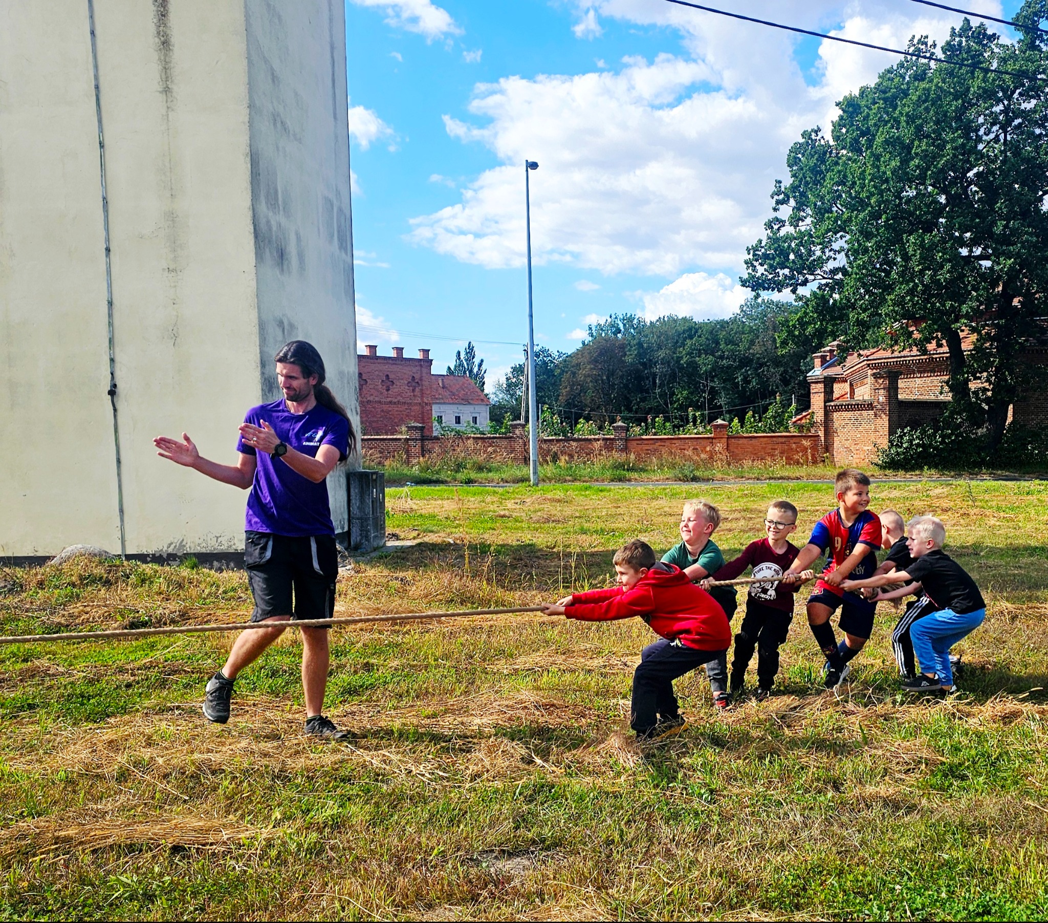 Animator w akcji: grupa dzieci uczestniczy w przeciąganiu liny na trawiastym terenie pod okiem mężczyzny w fioletowej koszulce. Zabawa na świeżym powietrzu, budynek w tle.