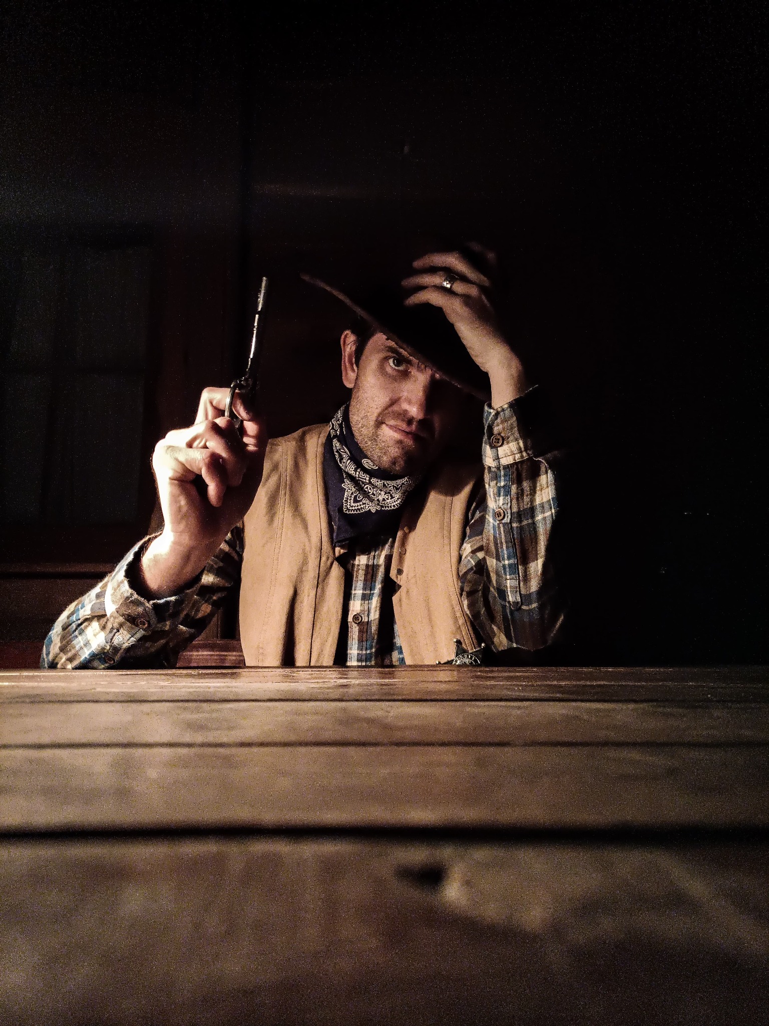 Mężczyzna w stroju kowboja, z chustą i kapeluszem, siedzi przy drewnianym stole i celuje z rewolweru. Mroczna sceneria, klimatyczny portret.