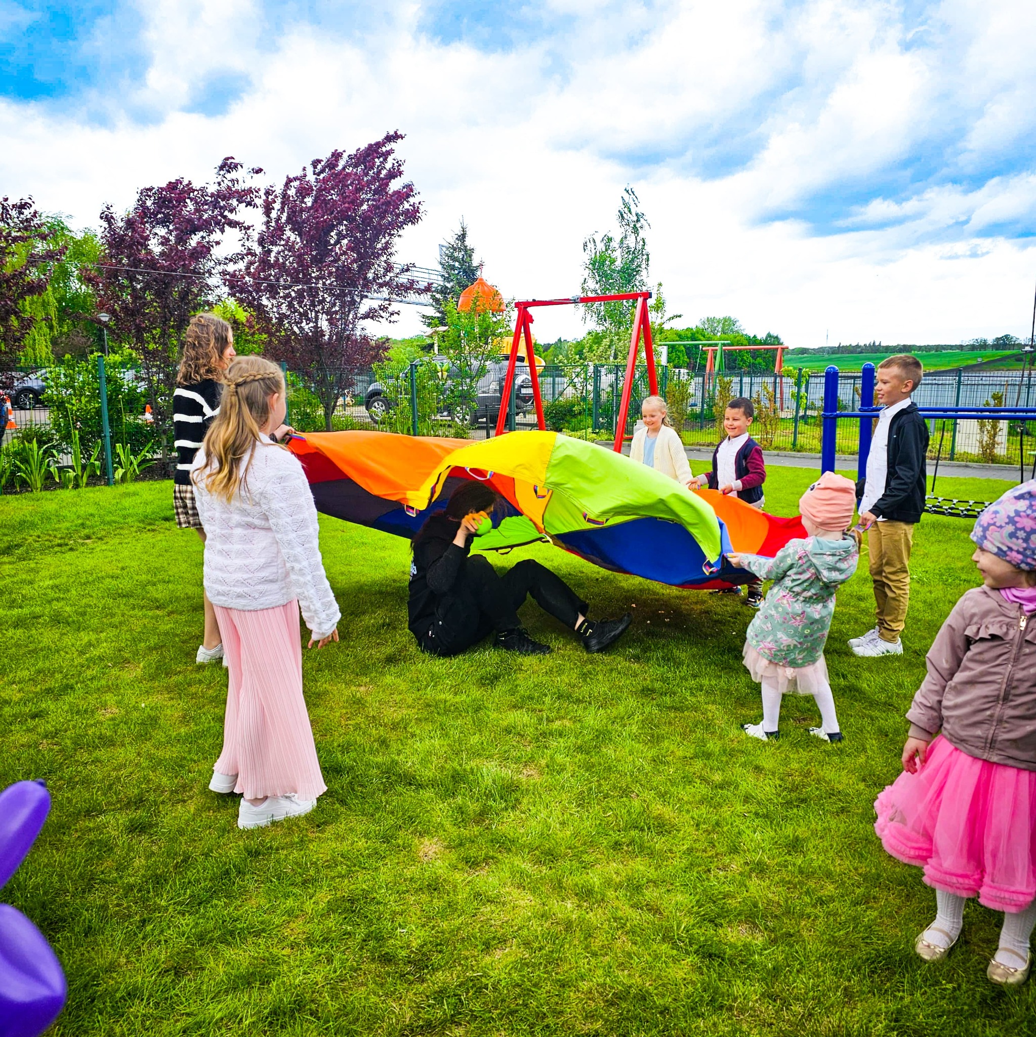 Animator bawi dzieci na zielonej trawie, używając kolorowej chusty animacyjnej. W tle plac zabaw i drzewa.