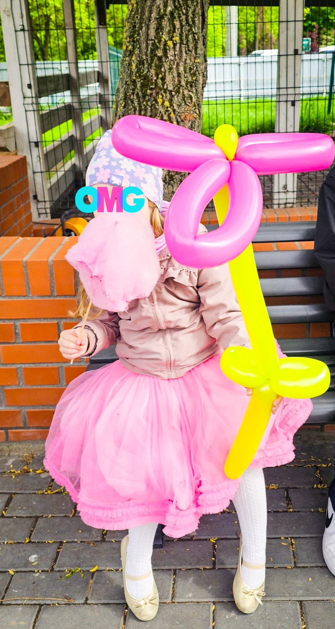 Dziewczynka w różowej tutu trzyma watę cukrową i balon w kształcie kwiatka na tle muru z cegły i zieleni.
